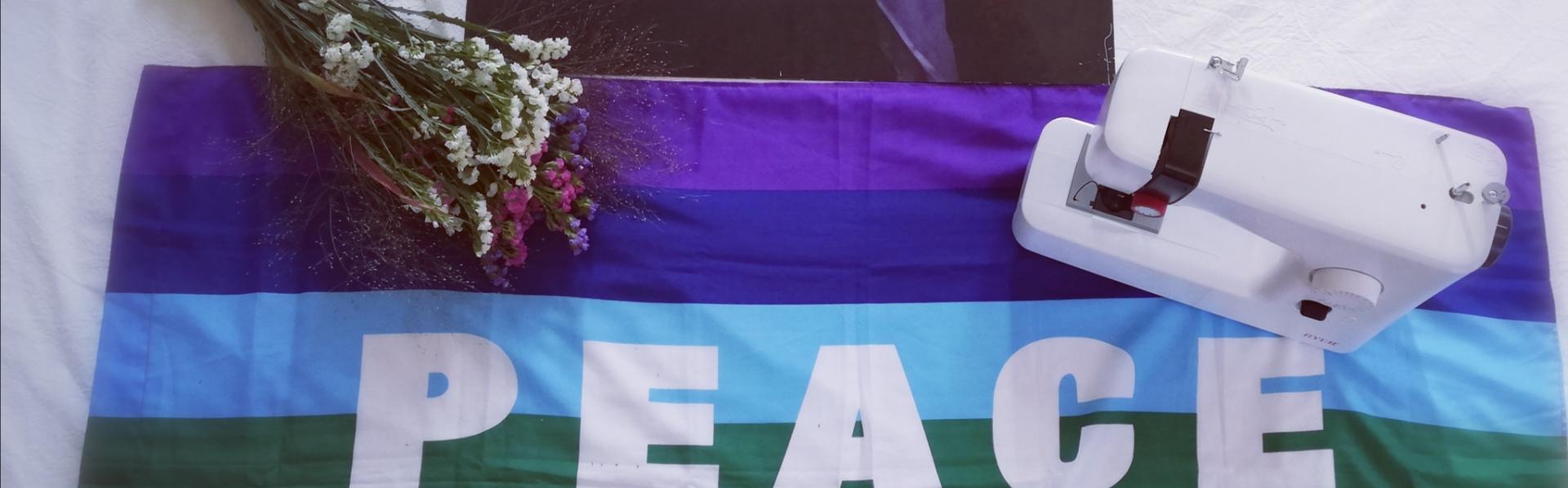 Vrouw met bloemen en doek boven een vredesvlag, geflankeerd door bosje gedroogde bloemen en een stikmachine.