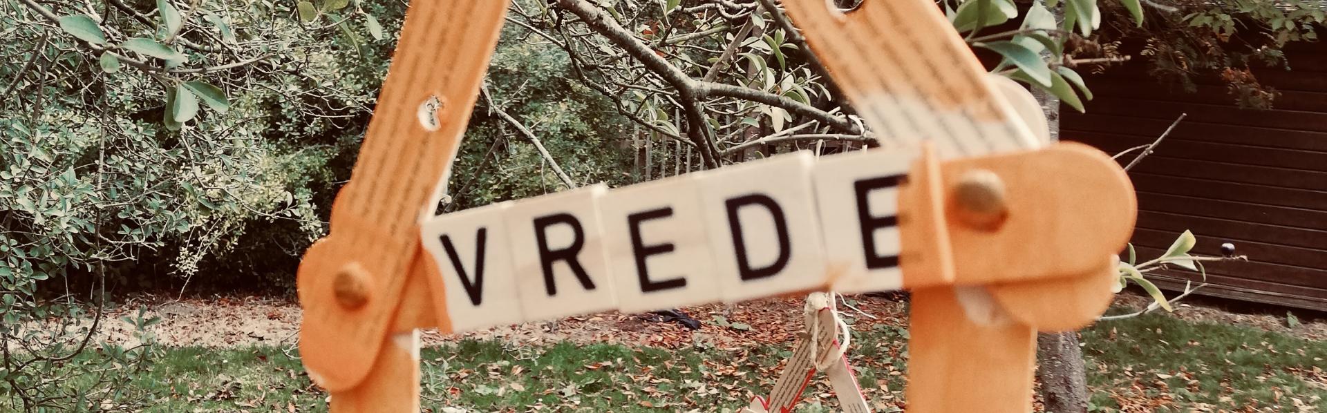 Een ééndimensionaal huisje van houten ijslollystokjes hangt buiten aan een tak. De dwarsbalk onder het dak leest VREDE.