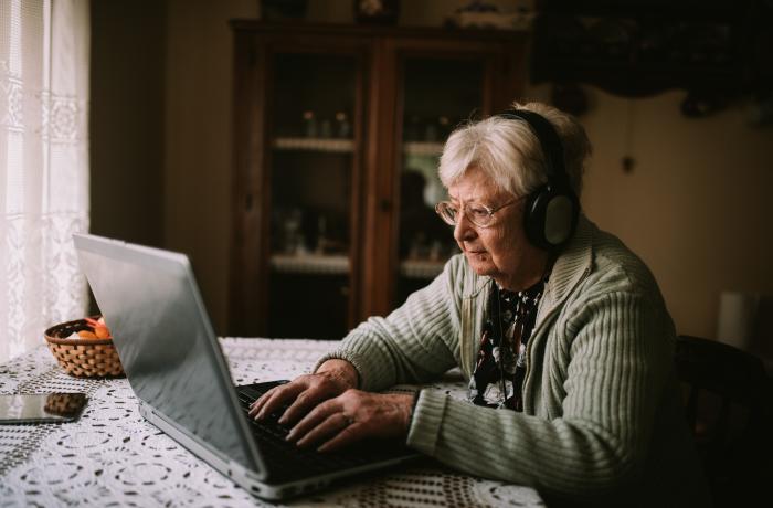 Oudere dame met hoofdtelefoon zit aan tafel, voor een laptop.