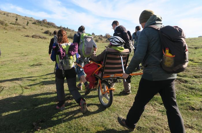 Een groep jongeren en ouderen samen aan het hiken. Eén iemand duwt en een andere trekt een rolkar met een man met helm in.