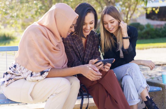 Drie glimlachende jonge diverse vrouwen kijken naar video op smartphone in park