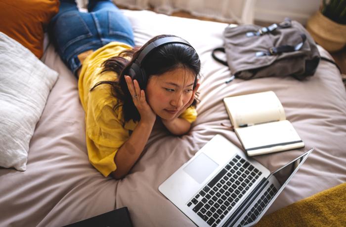 Jonge vrouw met hoofdtelefoons op bed, kijkt naar de computer. Een tas en schrift liggen naast haar.