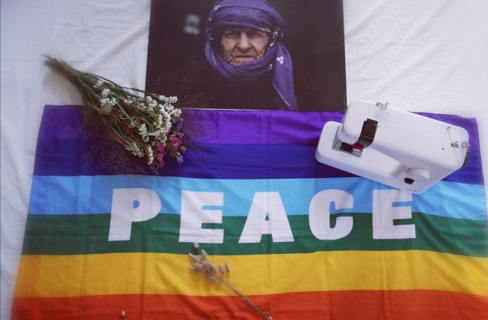 Vrouw met bloemen en doek boven een vredesvlag, geflankeerd door bosje gedroogde bloemen en een stikmachine.